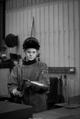 A woman in a welding helmet and glasses is using a blowtorch